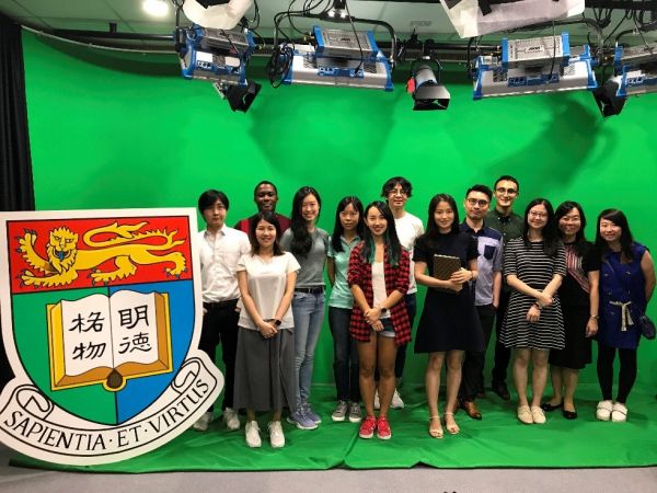 University of Hong Kong entrants standing as a group in front of a greenscreen for video creation next to large University logo